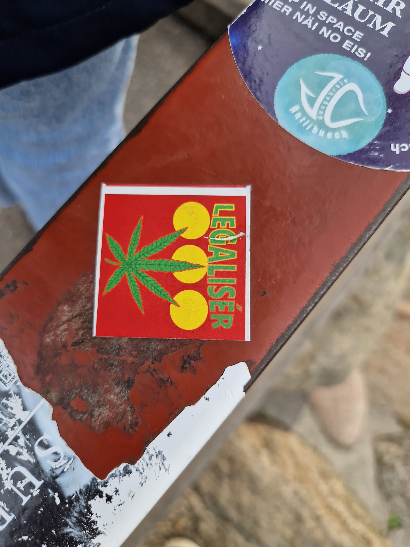 A red square sticker with a marijuana leaf and three yellow circles. The word "LEGALISER" is written vertically along the right side. The sticker is affixed to a rusty metal surface.