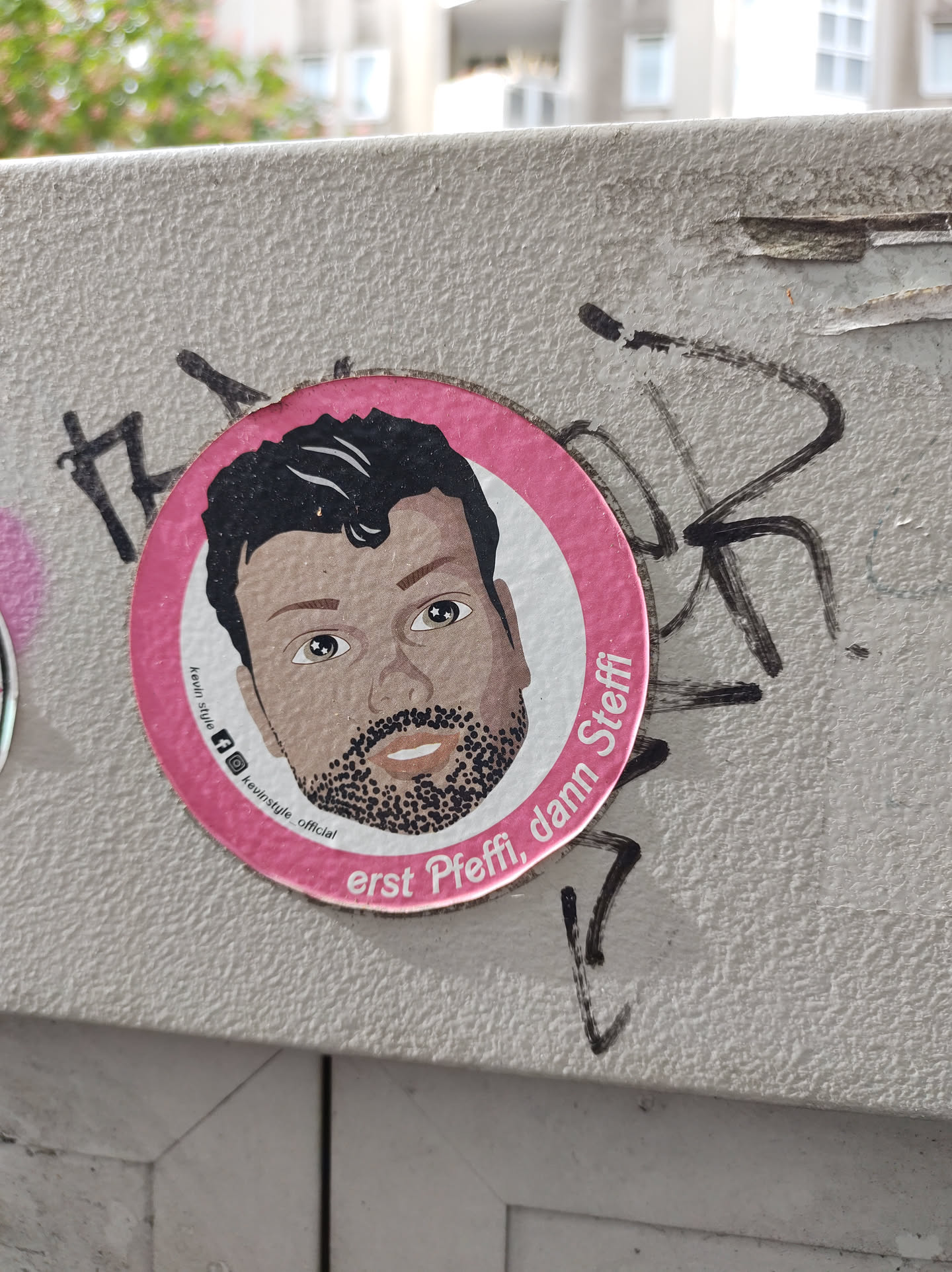 A round pink sticker with an illustration of a man's face. The text on the sticker says "erst Pffi, dann Steffi". There are some small icons below the illustration, including Instagram and maybe other social media accounts.