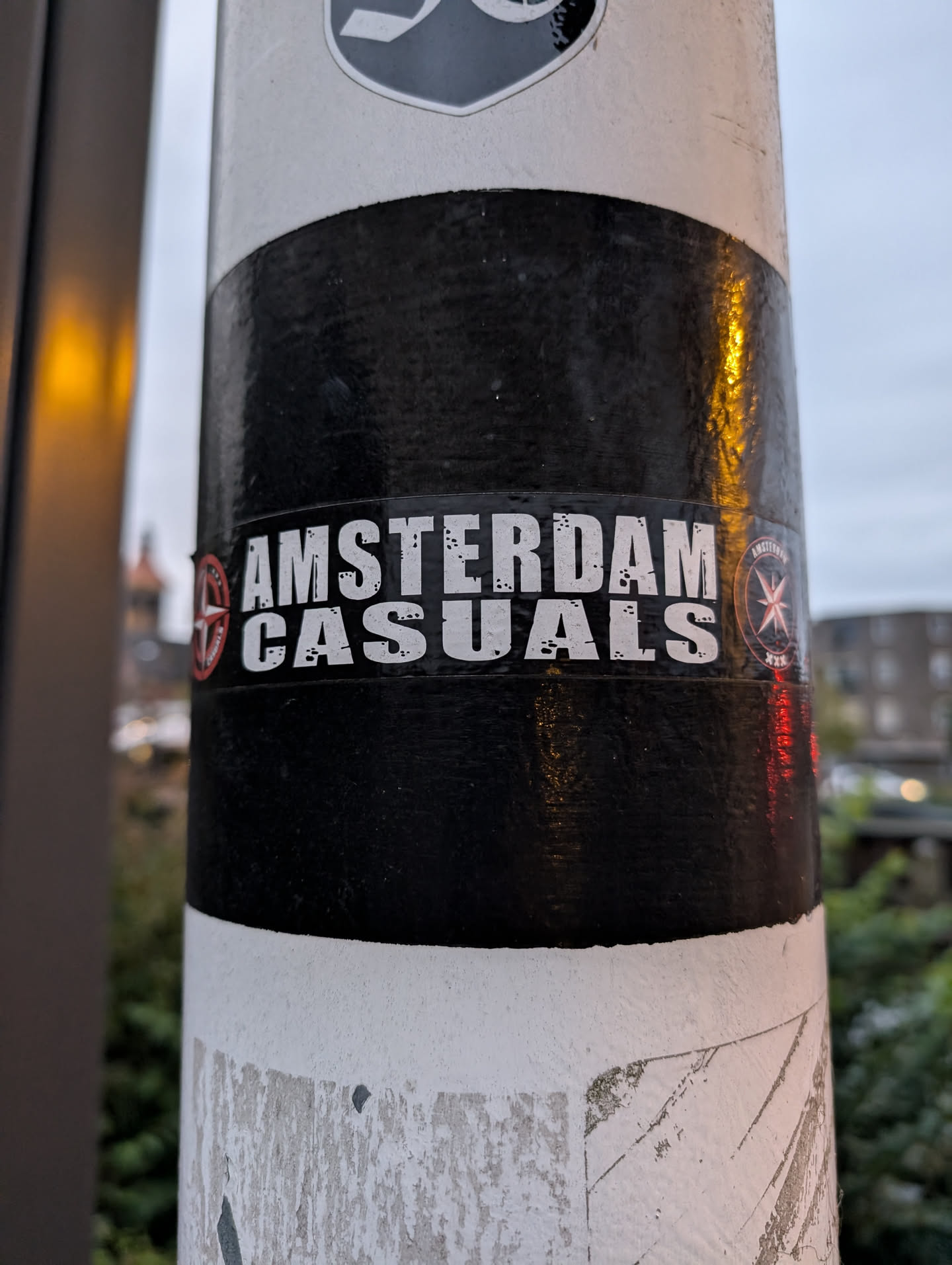 A black and white sticker with the text "AMSTERDAM CASUALS" in a bold, stylized font.  There are small circular symbols on either side of the text.
