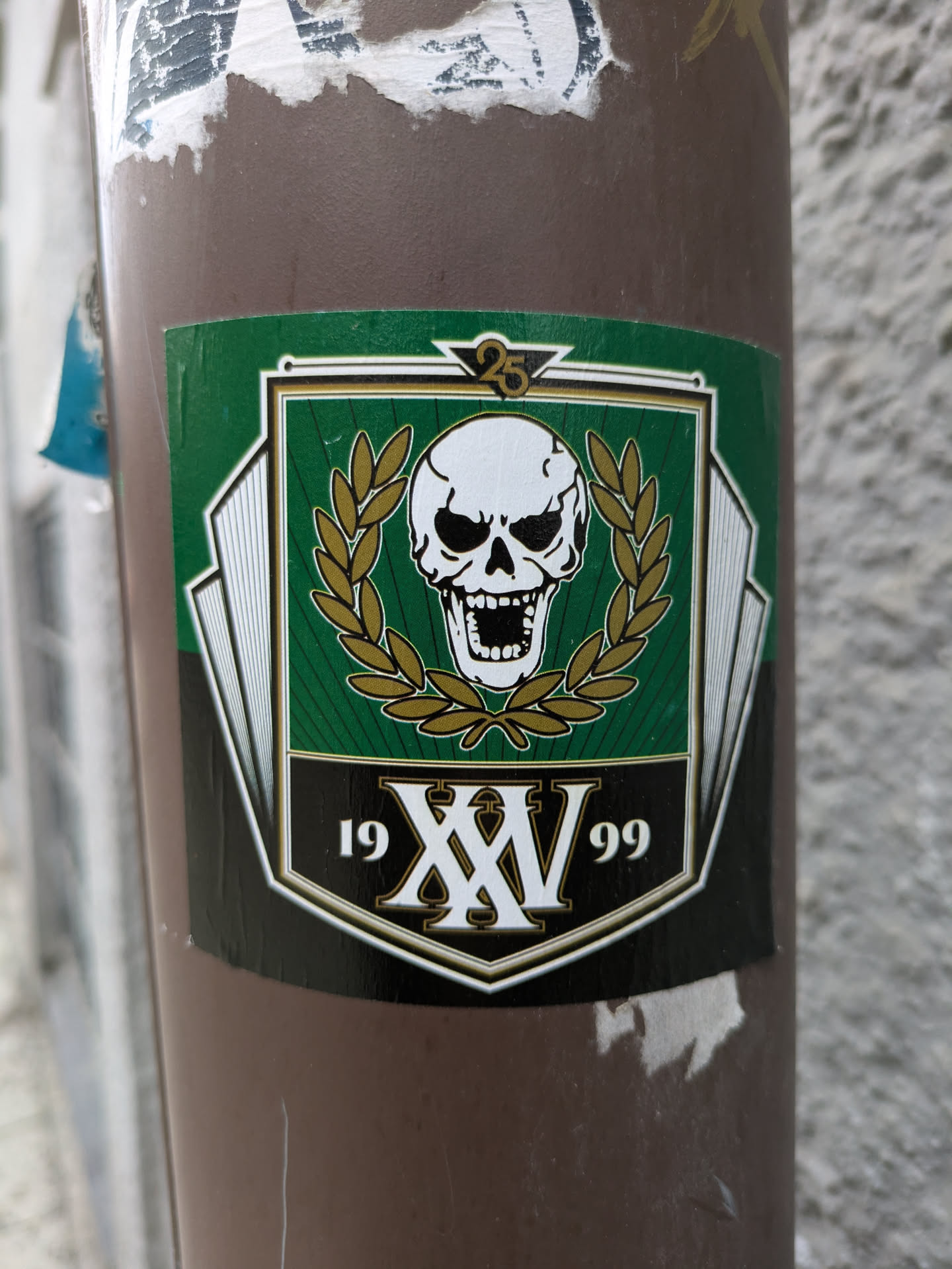 A sticker featuring a skull emblem with a laurel wreath, the roman numerals XV, and the years 19 and 99. It appears to commemorate a 25th anniversary.
