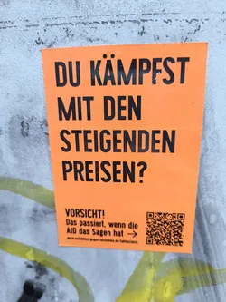An orange sticker affixed to a textured wall, asking 'DU KÄMPFST MIT DEN STEIGENDEN PREISEN?' ('Are you struggling with rising prices?'). The sticker also includes the word 'VORSICHT!' ('Caution!') followed by the phrase 'Das passiert, wenn die AfD das Sagen hat' ('This happens when the AfD has the say') and a QR code.