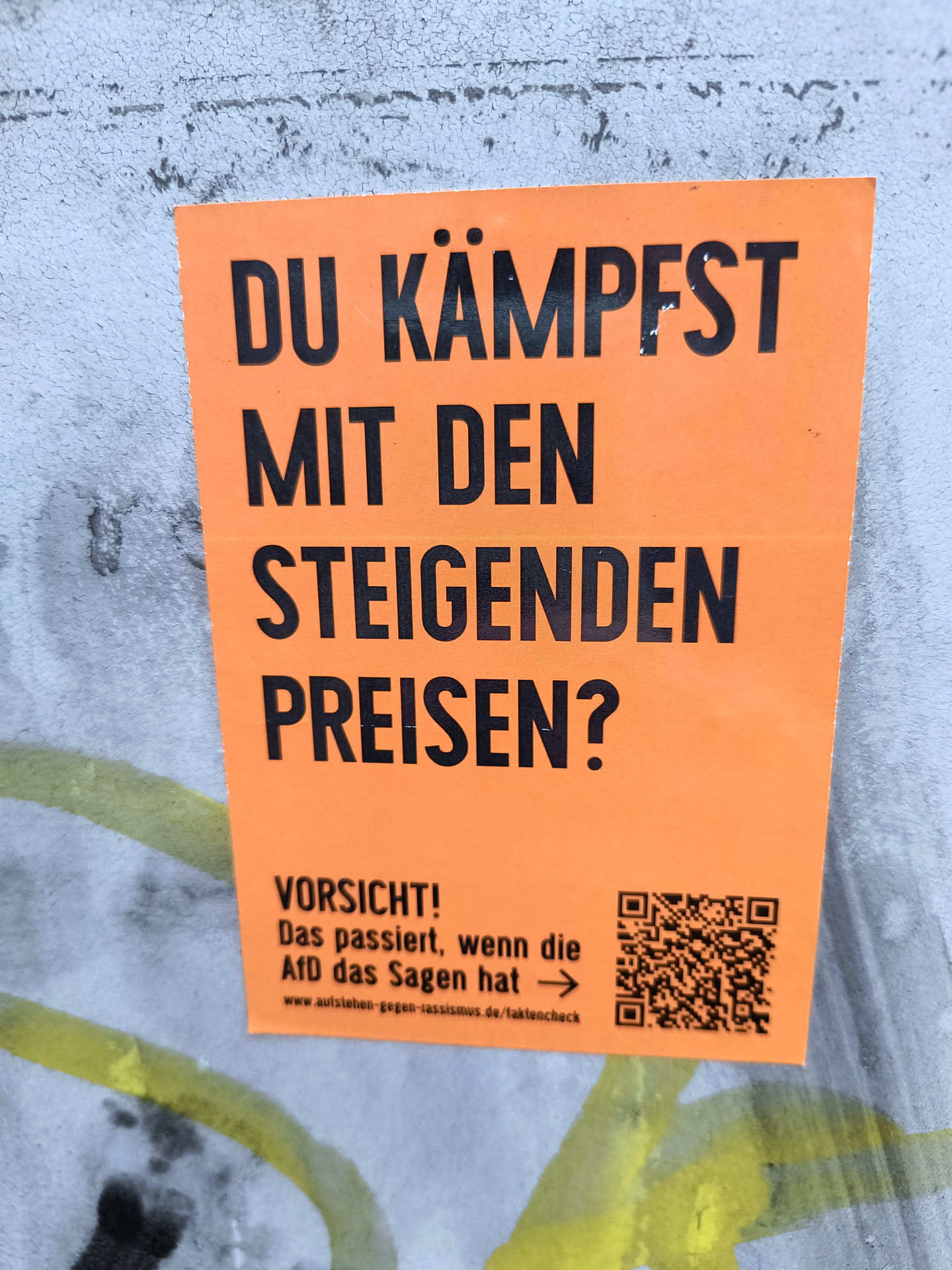 An orange sticker affixed to a textured wall, asking 'DU KÄMPFST MIT DEN STEIGENDEN PREISEN?' ('Are you struggling with rising prices?'). The sticker also includes the word 'VORSICHT!' ('Caution!') followed by the phrase 'Das passiert, wenn die AfD das Sagen hat' ('This happens when the AfD has the say') and a QR code.