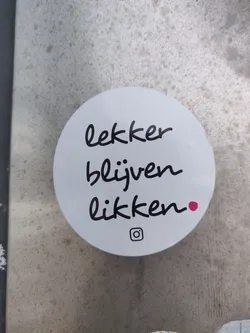A round white sticker with black handwritten text in Dutch that says "Lekker blijven likken."  It features a small pink dot and an Instagram logo.
