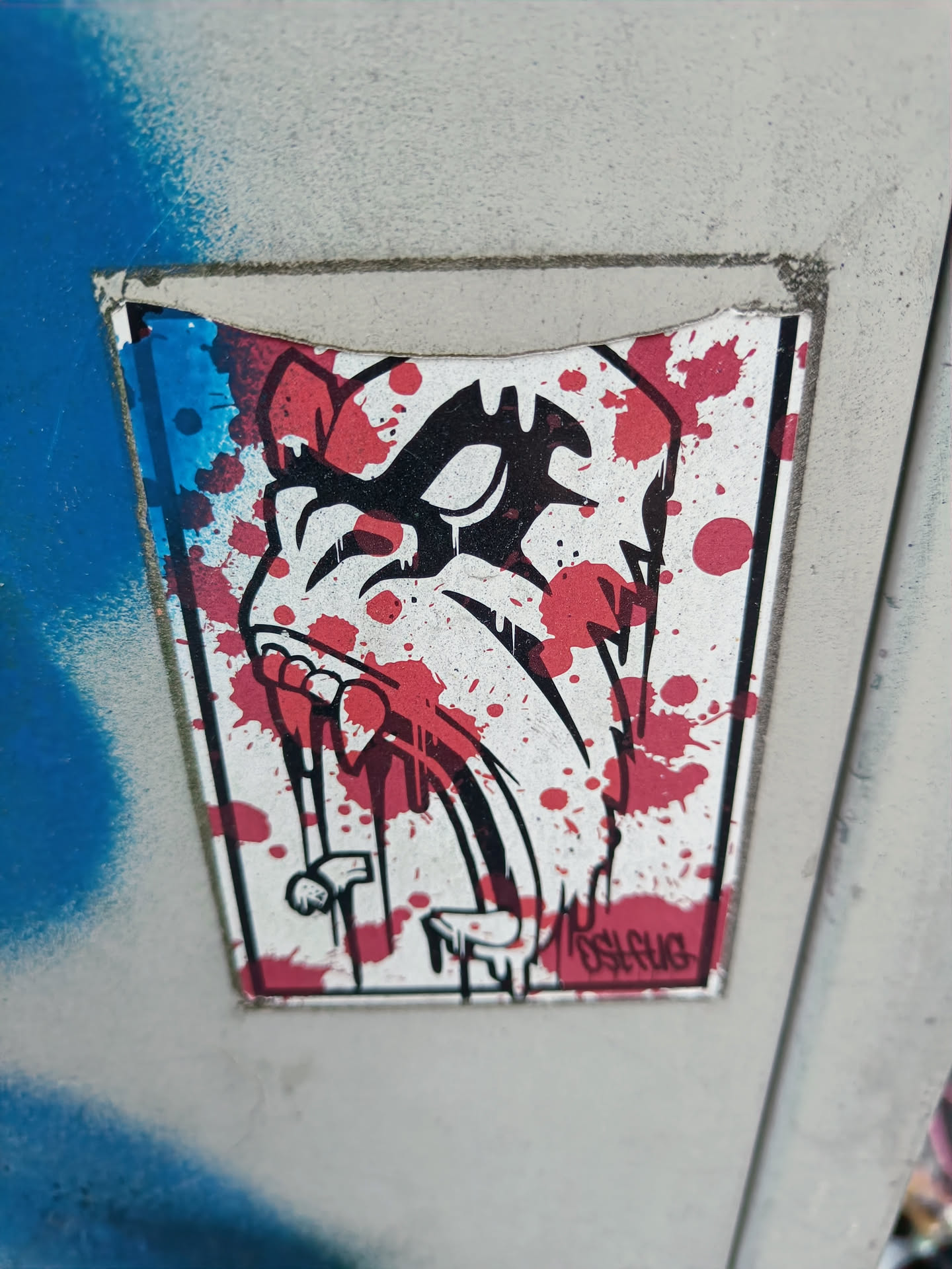 A sticker featuring a graphic illustration of a creature with sharp teeth and blood splatters. The style is reminiscent of horror or punk aesthetics. The artist's signature is visible in the bottom right corner.