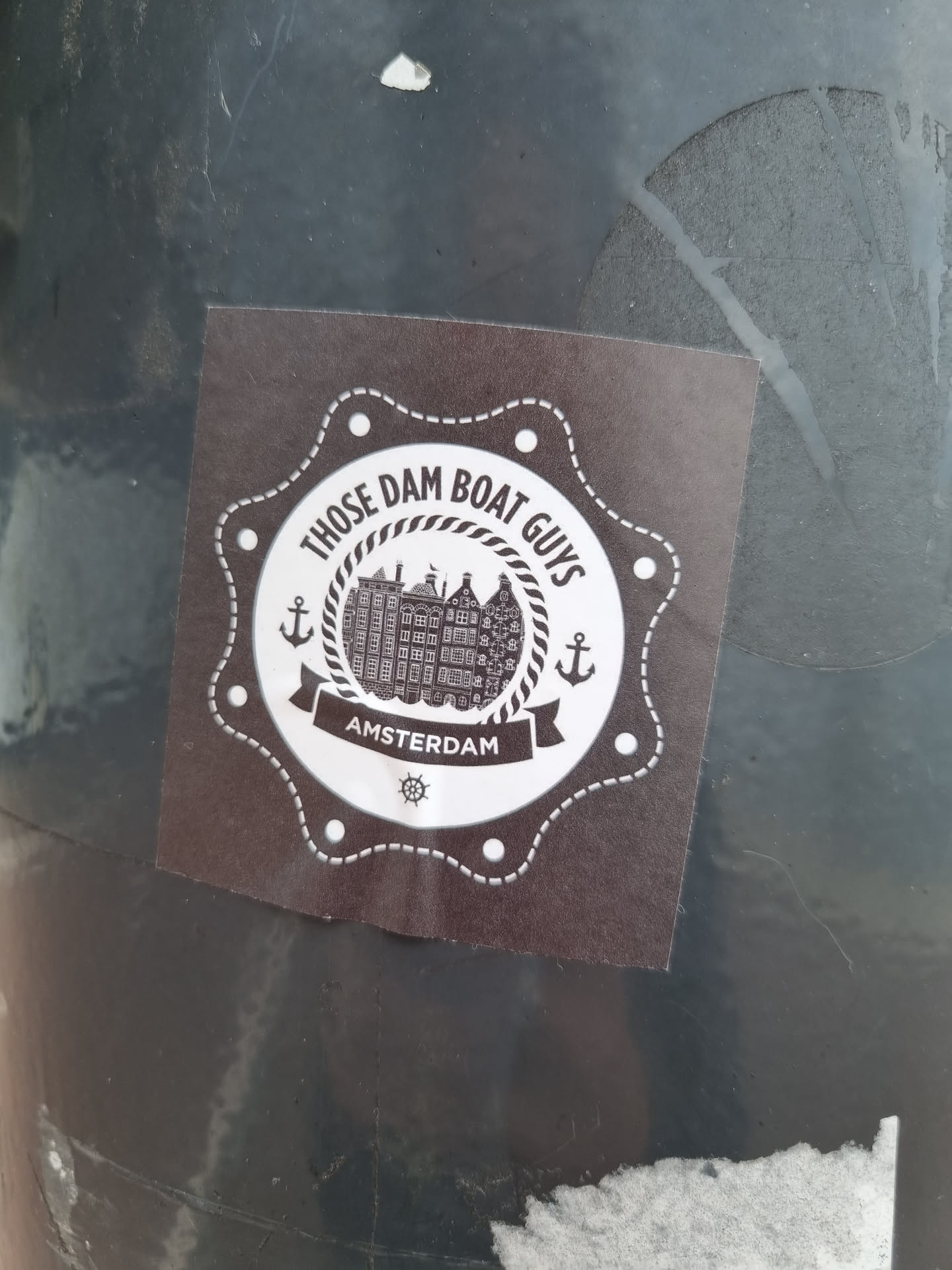 A dark-brown sticker with a circular design. It features the text "Those Dam Boat Guys" in a circular banner above an illustration of buildings, likely in Amsterdam.  "Amsterdam" is written beneath the illustration.  Anchors are displayed on each side of the circular banner. The sticker has a slightly distressed, vintage look.