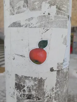 A round sticker featuring a realistic drawing of a red apple with a green leaf. The sticker is affixed to a white pole that shows signs of wear, with other, partially removed stickers visible in the background. 