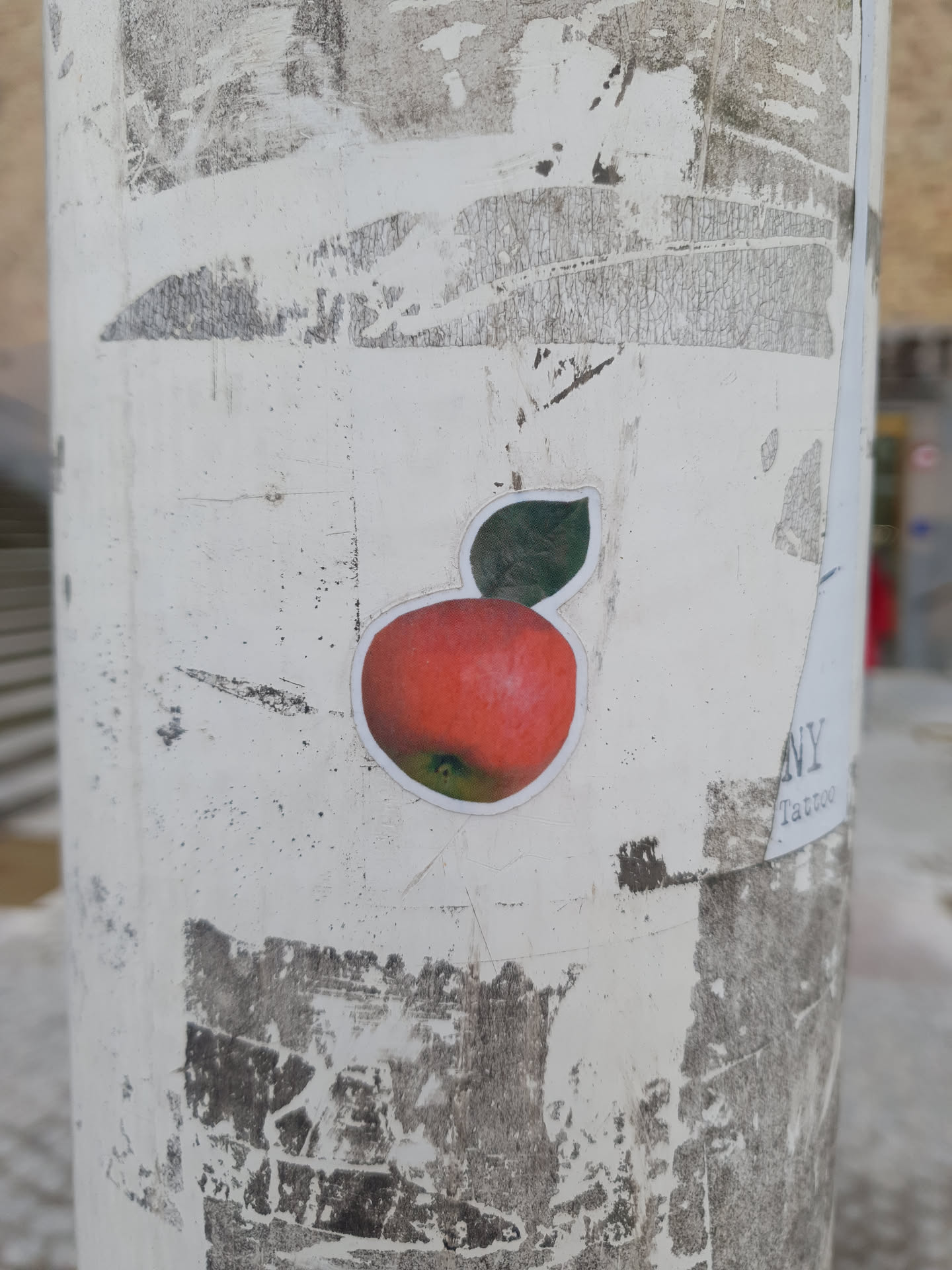A round sticker featuring a realistic drawing of a red apple with a green leaf. The sticker is affixed to a white pole that shows signs of wear, with other, partially removed stickers visible in the background. 