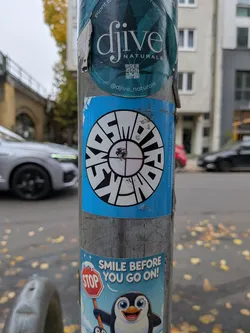 A rectangular, bright blue sticker attached to a metal pole. The center of the sticker features a circular emblem resembling a segmented lens or crosshair target. Warped within the segmented outer ring are bold black letters spelling out 'KOSMOTRONIKS' against white fills. In the core of the wheel is a four-quadrant target graphic with thin crosshair lines. Vertically along the right edge, the website address 'KOSMOTRONIKS.COM' is printed in clean, uppercase font.