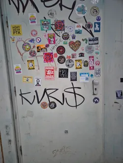 The image captures a light-colored, possibly off-white or pale green, metal door heavily covered with a dense and eclectic assortment of stickers and some black graffiti. The stickers vary significantly in size, shape (round, square, rectangular, die-cut), and artistic style, ranging from bold political statements to cartoonish illustrations, band logos, and humorous phrases. Prominent colors include bright reds, yellows, blues, greens, pinks, and black and white. Many stickers feature German text, indicating local activism and cultural references. Notable elements include a large yellow 'HÖRT MEHR PTK' sticker, a red 'GEGEN NAZIS' sticker, a heart-shaped sticker with a skull-like face, a 'FIGHT BORDERS' sticker, and a 'BERLINER KÄLTEHILFE' sticker with a QR code. Some stickers are peeling, faded, or partially obscured by other stickers or the prominent black graffiti 'KVRIS$' and 'SASAR'. The door itself shows signs of wear, dirt, and previous paint layers, contributing to an authentic urban aesthetic.
