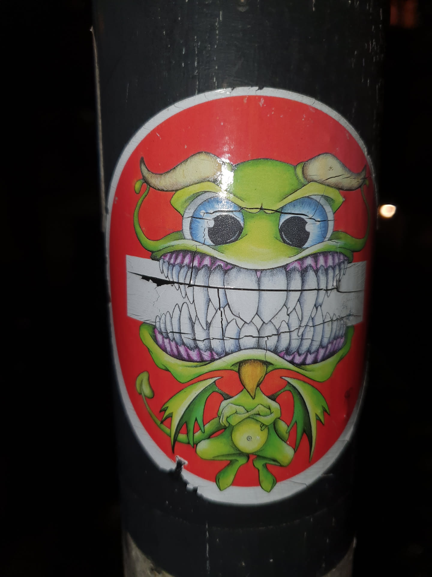 This sticker features a cartoonish green devil with large eyes, horns, and wings. It's got a big toothy grin, and its arms are crossed. The sticker has a red circle background and is slightly damaged around the edges.