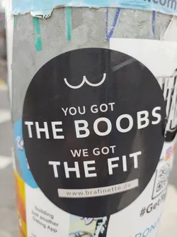 A black round sticker with white text on it. It says "YOU GOT THE BOOBS WE GOT THE FIT" and has a website address at the bottom: www.brafinette.de. There is a minimalist drawing of boobs at the top. 