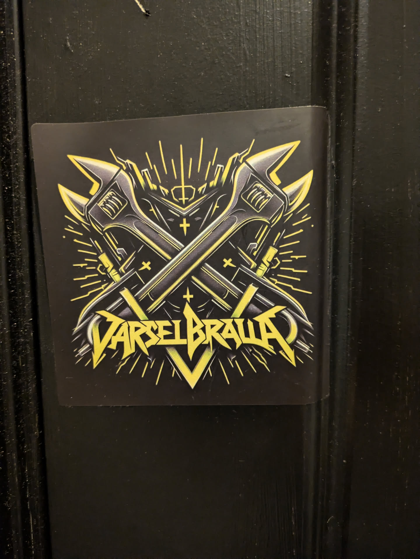 The sticker is predominantly black with vibrant yellow and metallic gray accents. At its center are two crossed adjustable wrenches, depicted with a metallic sheen. Behind and around the wrenches, stylized yellow lines radiate outwards, resembling bursts of energy or light. The overall design has a strong, industrial, and somewhat aggressive feel. Below the wrenches, bold, stylized yellow lettering spells out "VARSELBRAVA", with sharp angles and a metallic texture. Small cross symbols are subtly integrated into the design, adding to its symbolic nature.