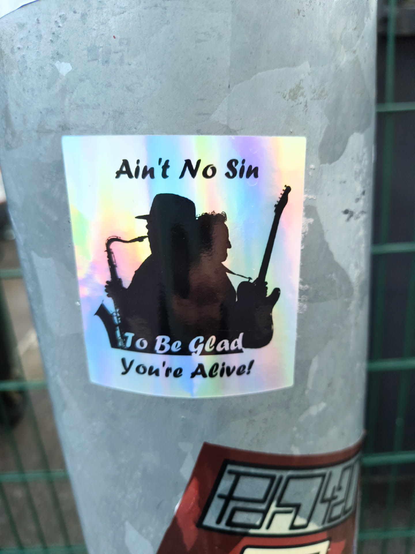 The sticker is a rectangular shape with slightly rounded corners, featuring a highly reflective, holographic background that shimmers with various pastel colors (blues, purples, pinks, yellows, greens) depending on the light angle. Against this vibrant backdrop, two prominent black silhouettes are depicted. On the left, a person wearing a hat is shown in profile, holding and playing a saxophone. On the right, another person, also in profile, is playing an electric guitar. Both figures appear to be musicians. Above the silhouettes, the text "Ain't No Sin" is written in black, bold, slightly italicized font. Below the silhouettes, more text reads "To Be Glad" on one line and "You're Alive!" on the line below it, in the same font style. The sticker is applied to a textured, light grey metallic pole.