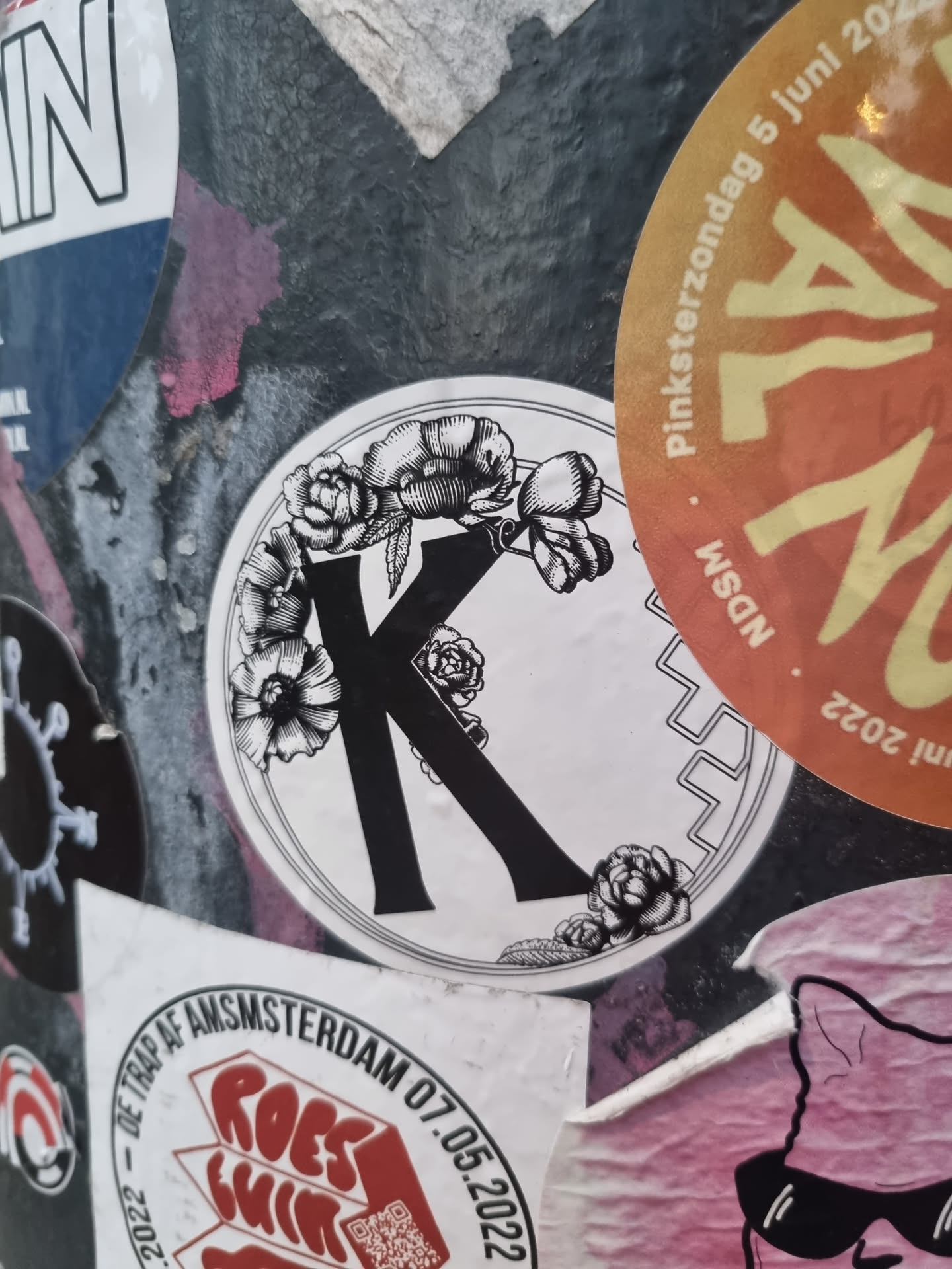 A round sticker featuring a stylized letter K in black, adorned with detailed floral illustrations in the same color scheme. The background is white, creating a striking contrast. The style suggests a vintage or botanical aesthetic.