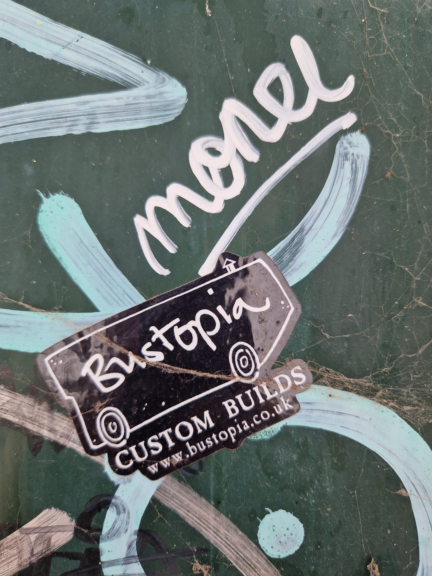 Black and white sticker with the text 'Bustopia' and 'CUSTOM BUILDS' with a website address. The sticker is placed on a green surface with white graffiti.