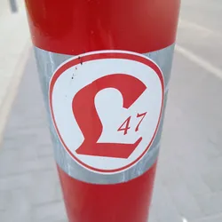 A close-up image of a round sticker affixed to a red pole. The sticker features a stylized £ symbol with the number 47 underneath, all in red against a white background.  The sticker shows some minor wear and discoloration.