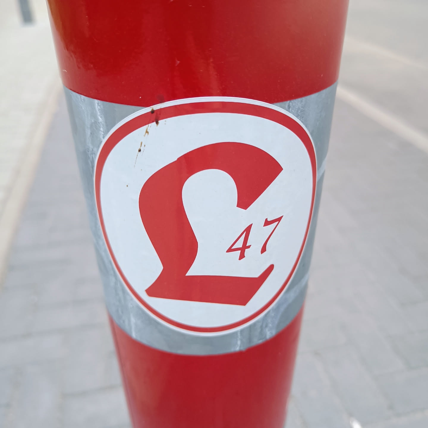 A close-up image of a round sticker affixed to a red pole. The sticker features a stylized £ symbol with the number 47 underneath, all in red against a white background.  The sticker shows some minor wear and discoloration.