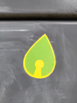This sticker features a teardrop shape with a yellow keyhole symbol inside.  The teardrop is green, and the keyhole is a bright yellow. The sticker is affixed to a dark gray surface.