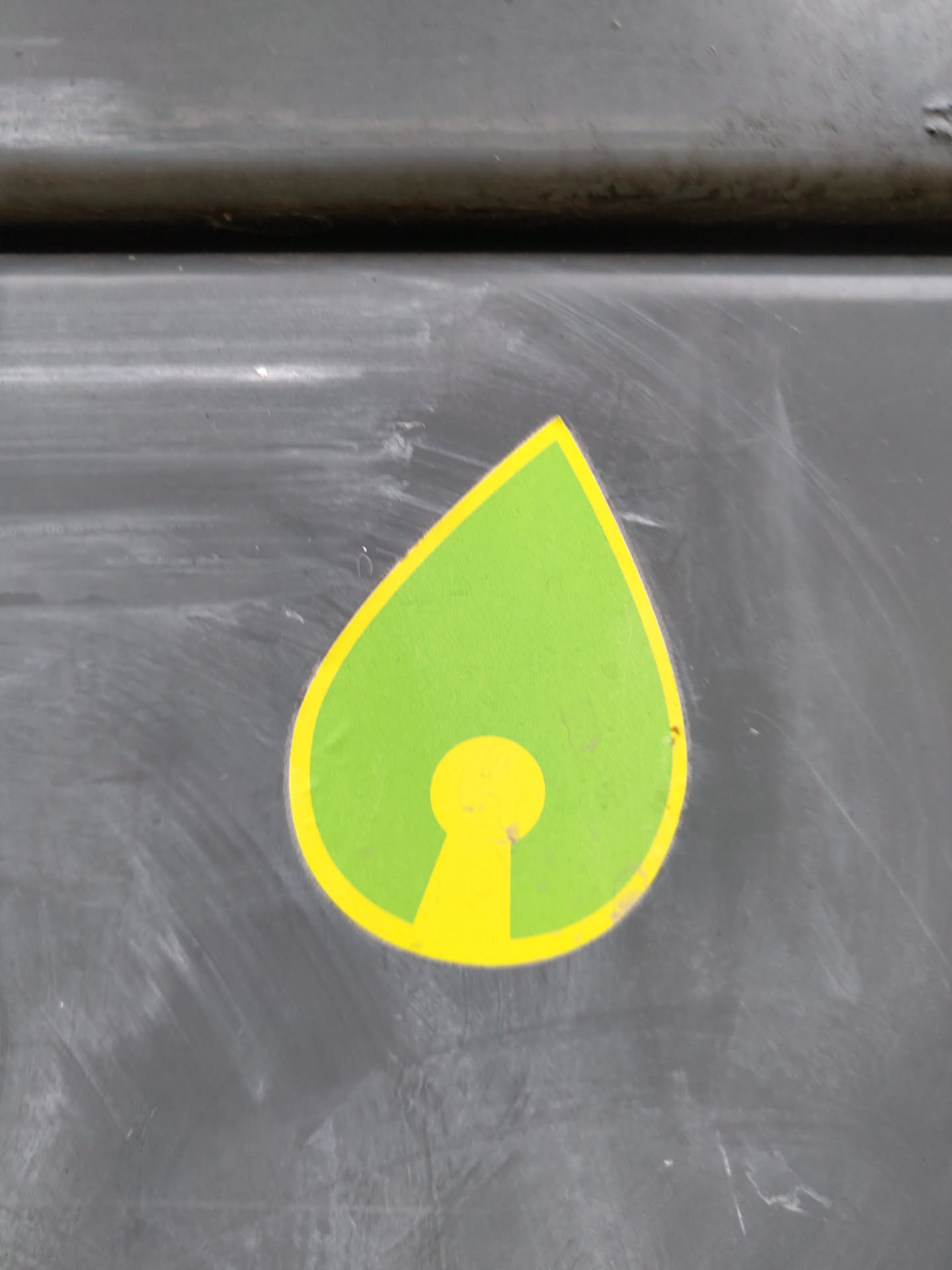 This sticker features a teardrop shape with a yellow keyhole symbol inside.  The teardrop is green, and the keyhole is a bright yellow. The sticker is affixed to a dark gray surface.