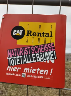 A sticker reading "Natur ist Scheiße Tötet alle Bäume!" (Nature is shit, kills all trees!) is affixed to a Cat Rental Store sign.  The sticker is in German and expresses environmental protest.