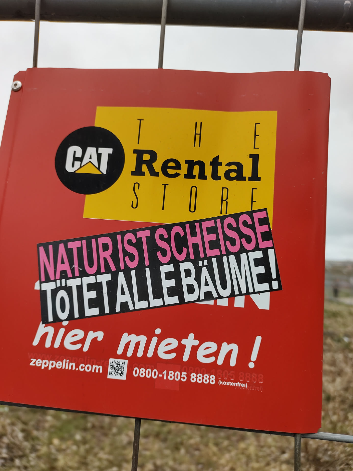 A sticker reading "Natur ist Scheiße Tötet alle Bäume!" (Nature is shit, kills all trees!) is affixed to a Cat Rental Store sign.  The sticker is in German and expresses environmental protest.