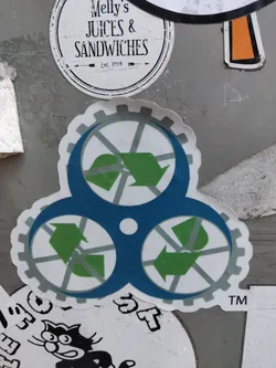 A sticker featuring a circular economy symbol. The symbol depicts three interconnected gears with green arrows indicating a circular flow. The gears are teal with gray accents. The sticker has a trademark symbol (TM) in the bottom right corner.