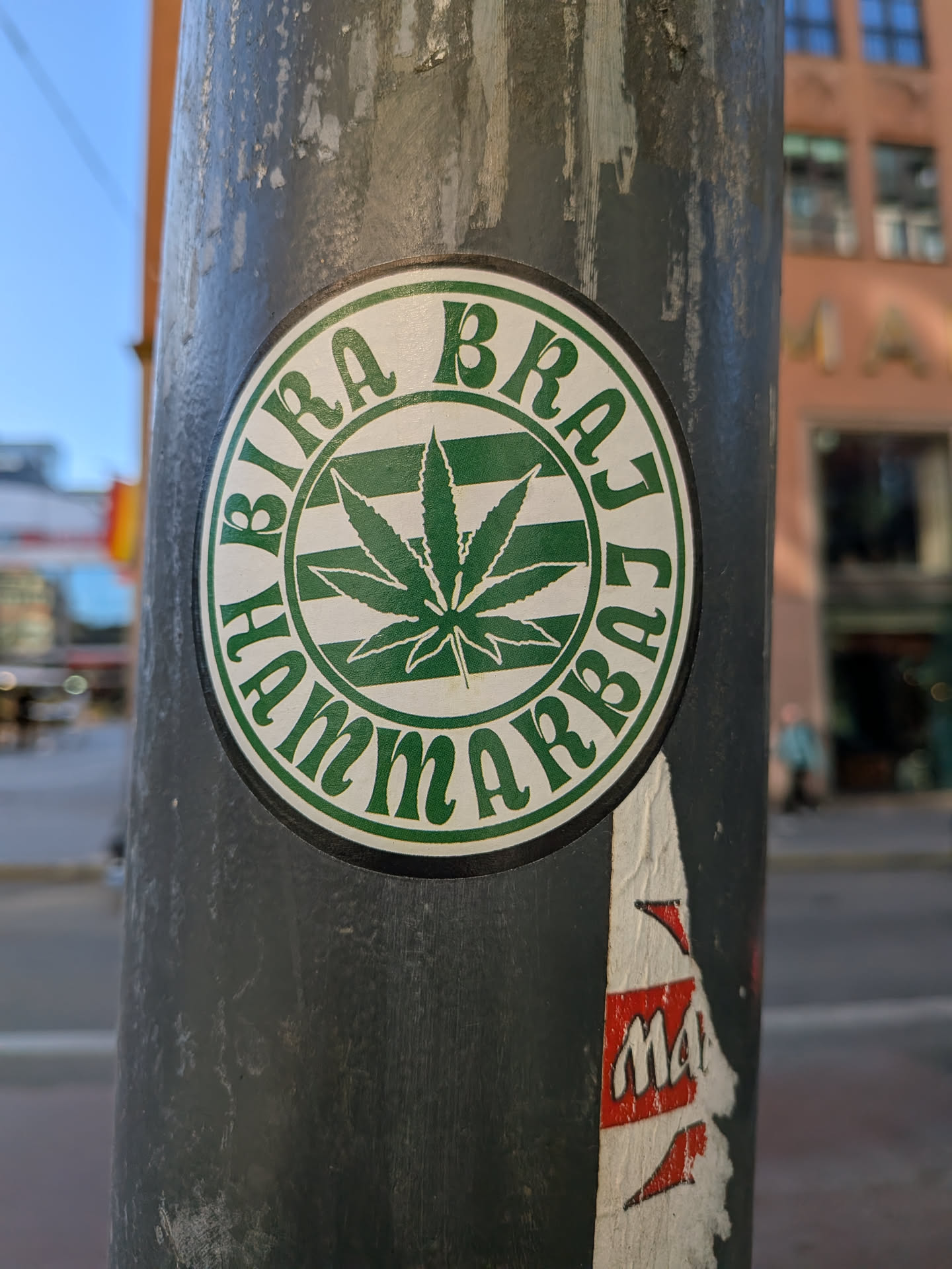 The sticker is circular, primarily green and white. At its center is a prominent, stylized green cannabis leaf, set against a white background with horizontal green stripes. Encircling this central design, along the outer edge of the sticker, is the text 'BIRA BRAJ HAMMARBY' written in green capital letters on a white background. The entire emblem is framed by a thin green border. The sticker shows some signs of wear and is attached to a dark, weathered utility pole.