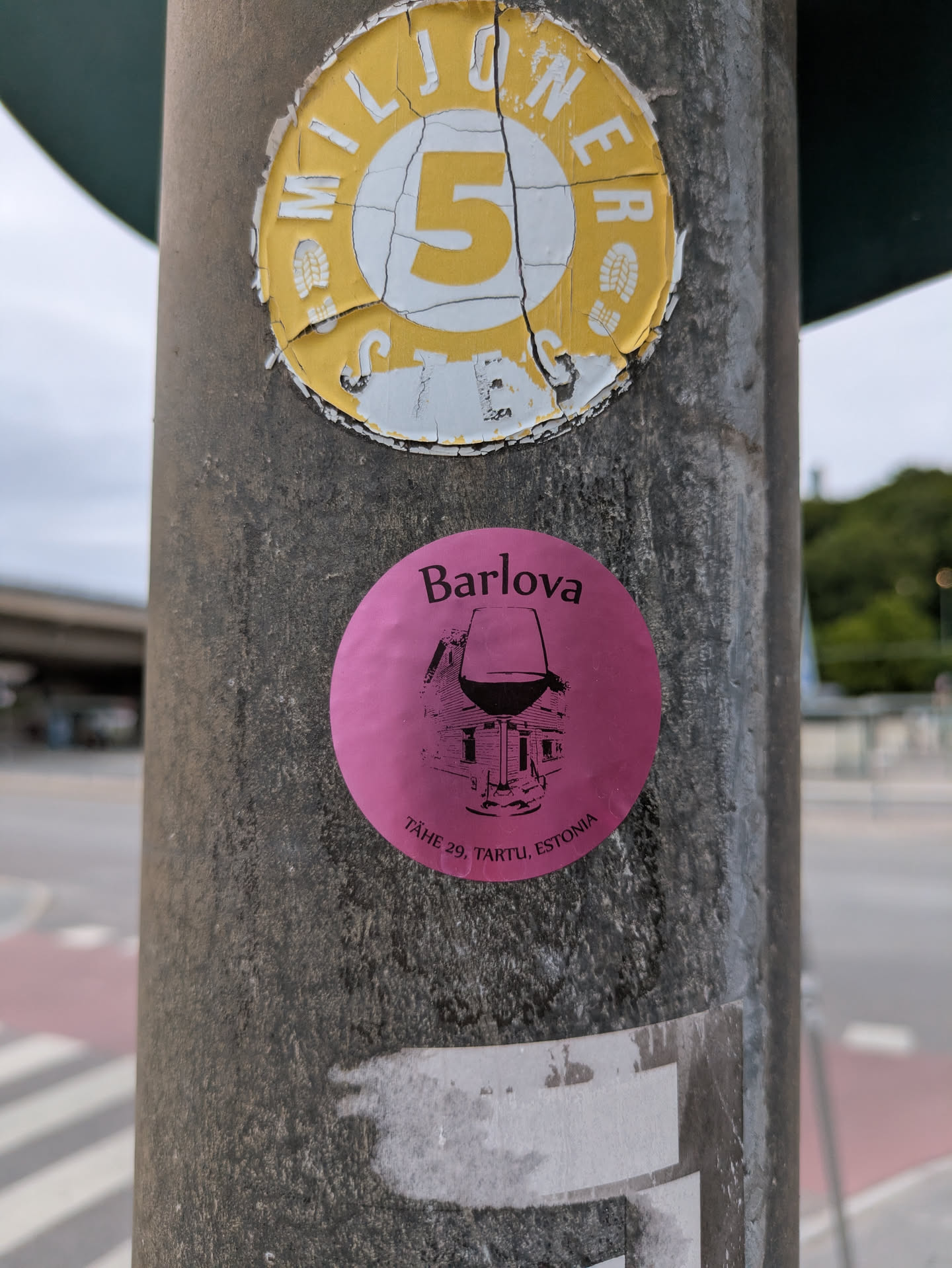 The central sticker is circular and bright pink (magenta). At the top, the word "Barlova" is written in black text. Below it, there is an intricate black line drawing of a wine glass, within the base and stem of which is depicted a detailed illustration of a building or house. Along the bottom curve of the sticker, the address "TÄHE 29, TARTU, ESTONIA" is printed, indicating a specific location in Tartu, Estonia. The sticker is affixed to a weathered grey pole.
