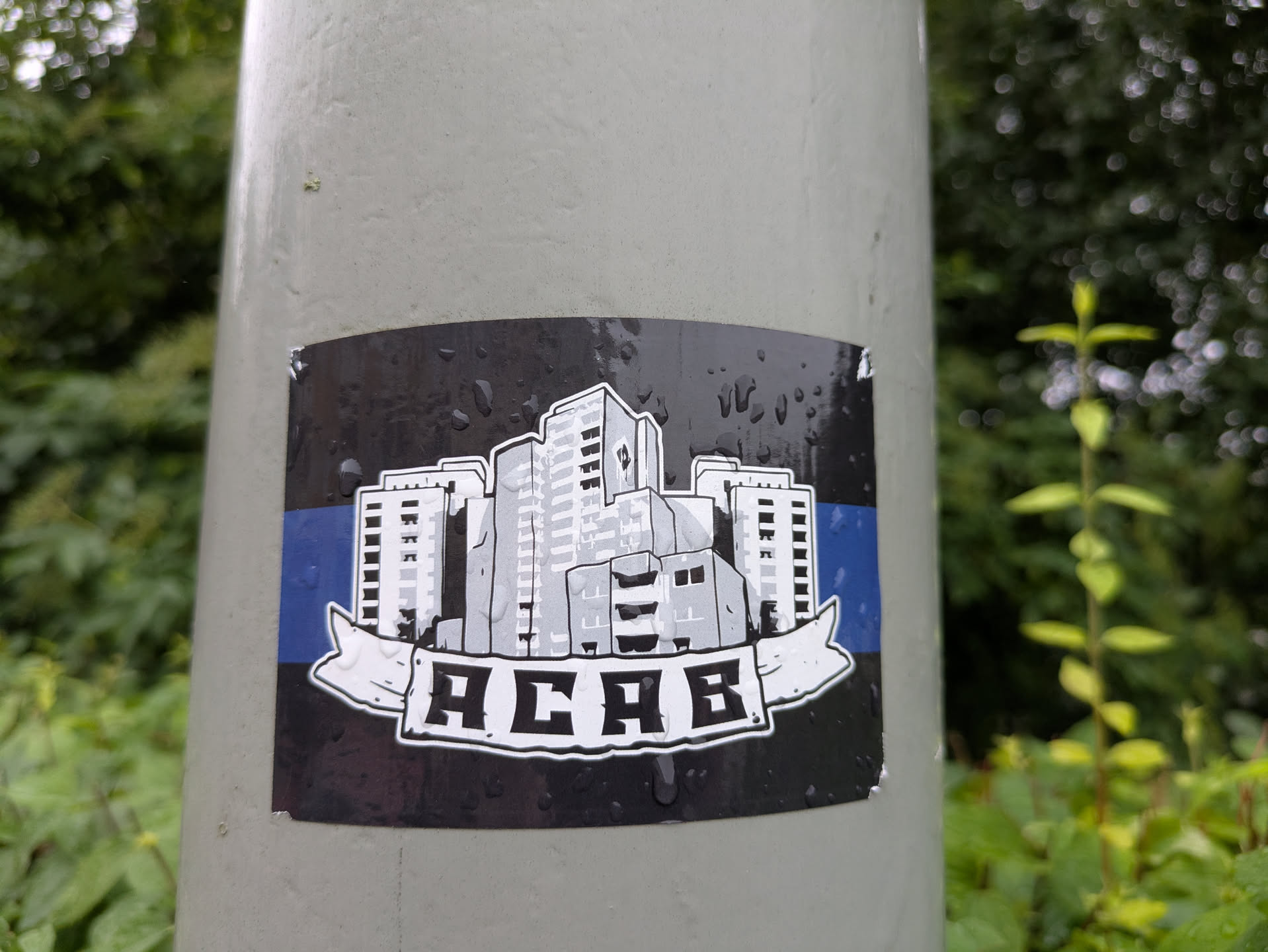 The sticker is rectangular with slightly rounded corners, adhered to a light grey pole. Its background is dark grey or black, covered with numerous small water droplets. Across the center, a horizontal blue stripe is visible. Overlaid on this stripe and the dark background is an illustration of a cluster of buildings in varying shades of grey and white, giving a monochrome, almost sketched appearance. These buildings are stylized, resembling apartment blocks or high-rises. Below the cityscape, a white banner with jagged ends unfurls, containing the bold, blocky text "ACAB" in black. The entire illustration (buildings and banner) has a white outline, making it stand out from the dark background. The sticker appears slightly worn at the top right corner.