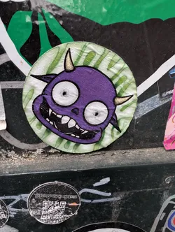 The sticker features a purple cartoon demon with small horns, wide eyes, and a toothy grin. It is circular with a green striped background.