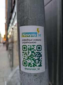 A sticker for Novyny.se, a Swedish news source in Ukrainian, is affixed to a pole. The sticker features a QR code and their Telegram handle.