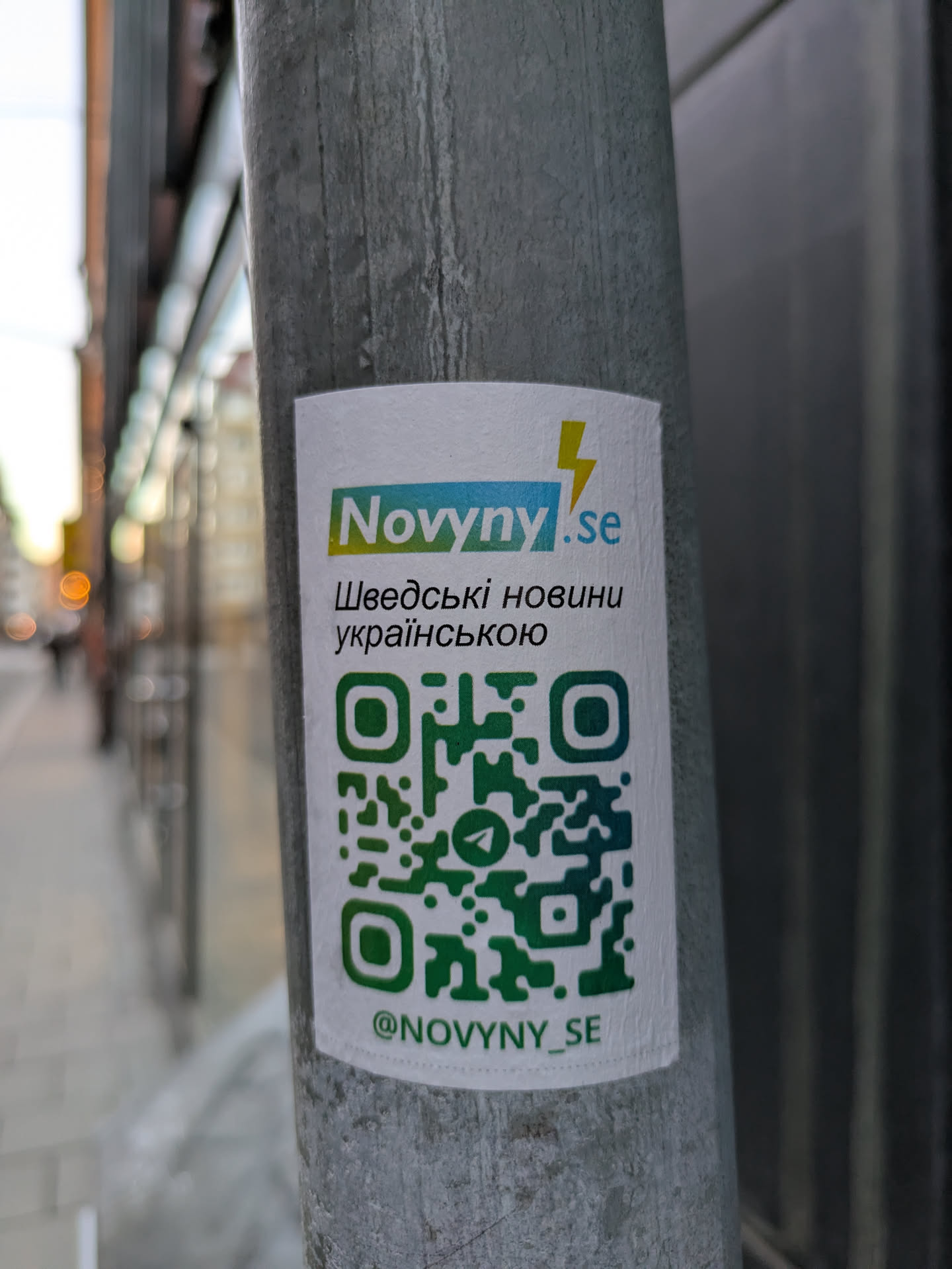 A sticker for Novyny.se, a Swedish news source in Ukrainian, is affixed to a pole. The sticker features a QR code and their Telegram handle.