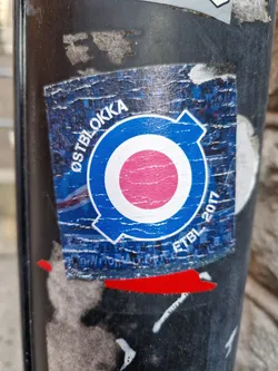 A worn sticker with a blue circle containing a pink circle in the center and the text "OSTBLOKKA" and "ETBL 2017". The sticker shows signs of wear and tear, with parts of it peeling off.