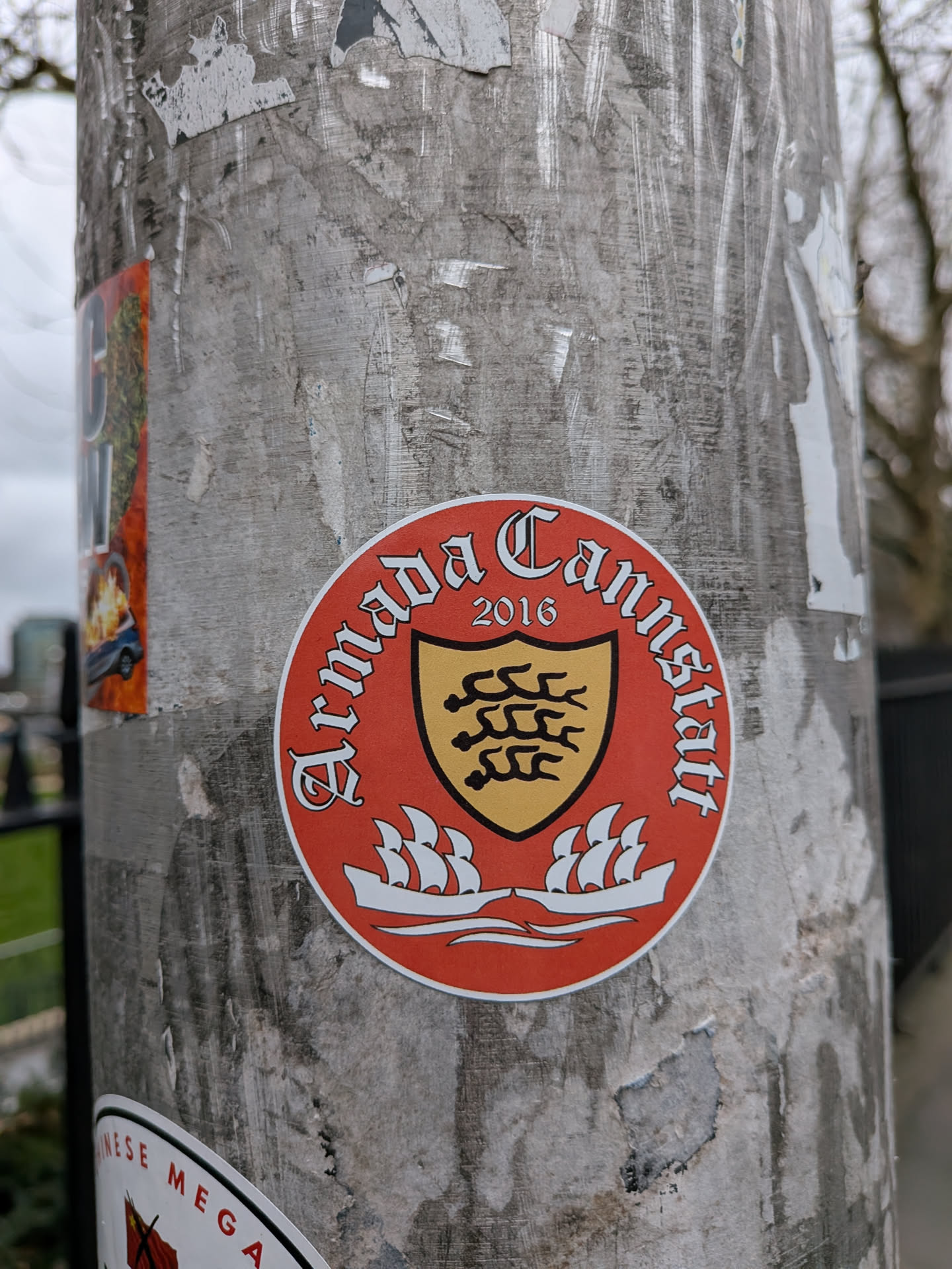 A circular sticker featuring a vibrant red outer ring with white text 'Armada Cannstatt' curved along the top and '2016' below it. The center of the sticker displays a gold shield outlined in black, containing a black design resembling three stylized antlers or waves, characteristic of the Württemberg coat of arms. Below the shield, two white sailing ships with multiple sails are depicted on white waves, all set against the red background of the sticker's lower half. The overall design evokes a sense of historical or group identity.