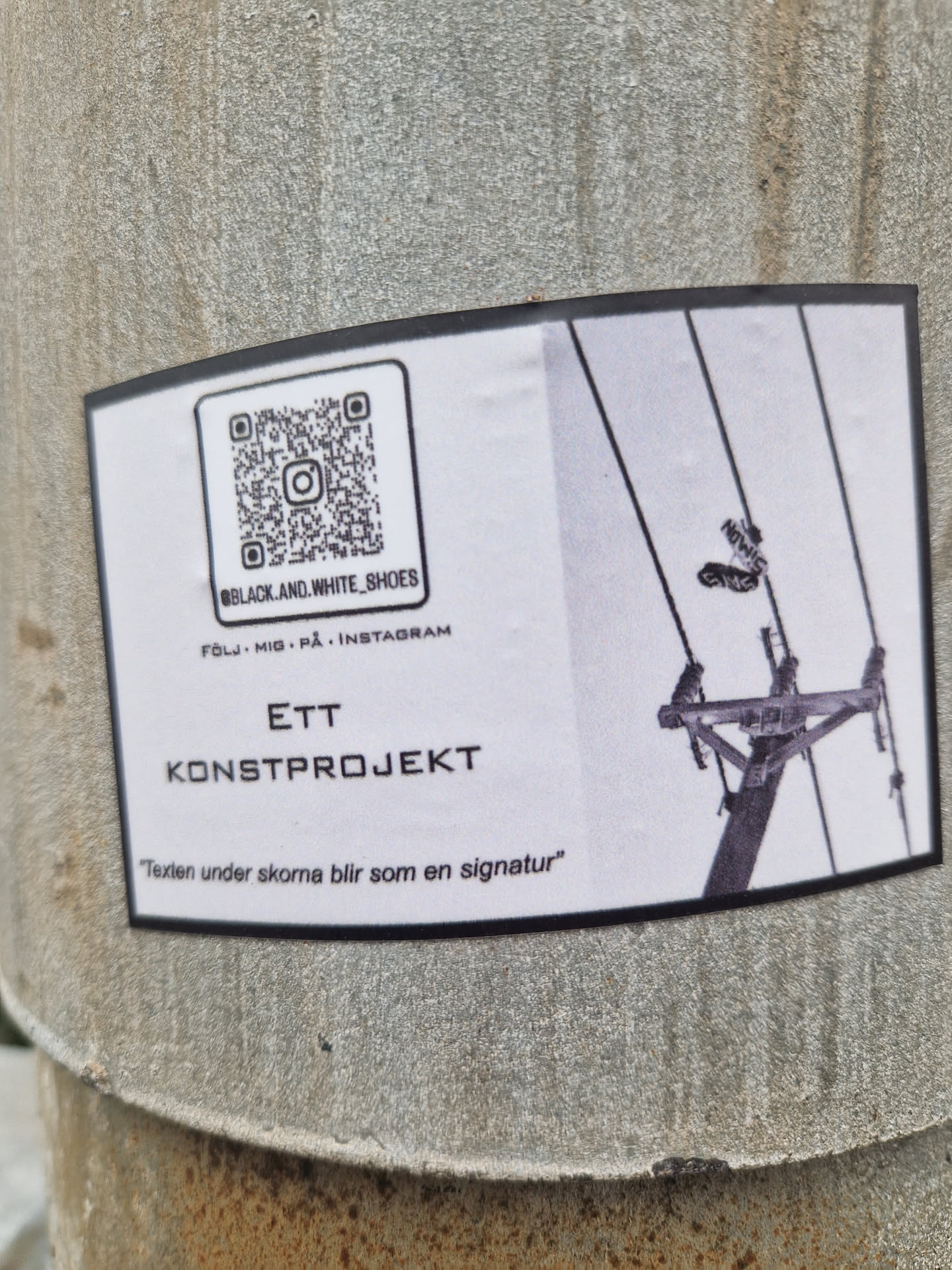 The sticker is rectangular with rounded corners, primarily white with a black border, adhered to a textured grey concrete surface. The left side features a QR code and the Instagram handle "@BLACK.AND.WHITE_SHOES" with the Swedish text "FÖLJ MIG . PÅ . INSTAGRAM" (Follow me . On . Instagram) underneath. Below this, the text "ETT KONSTPROJEKT" (An Art Project) is displayed prominently, followed by a quote: ""Texten under skorna blir som en signatur"" (The text under the shoes becomes like a signature). The right side of the sticker presents a black and white photograph depicting a utility pole with power lines, from which two objects resembling shoes are suspended.