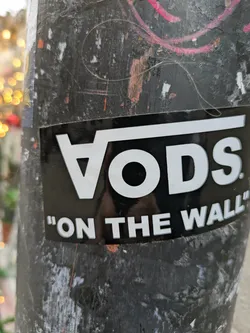 A black and white sticker with the word "Vods" in a stylized font and the phrase "On The Wall" underneath. The sticker is affixed to a weathered pole.