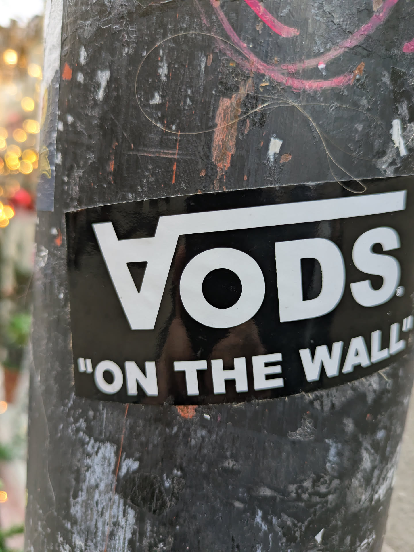 A black and white sticker with the word "Vods" in a stylized font and the phrase "On The Wall" underneath. The sticker is affixed to a weathered pole.
