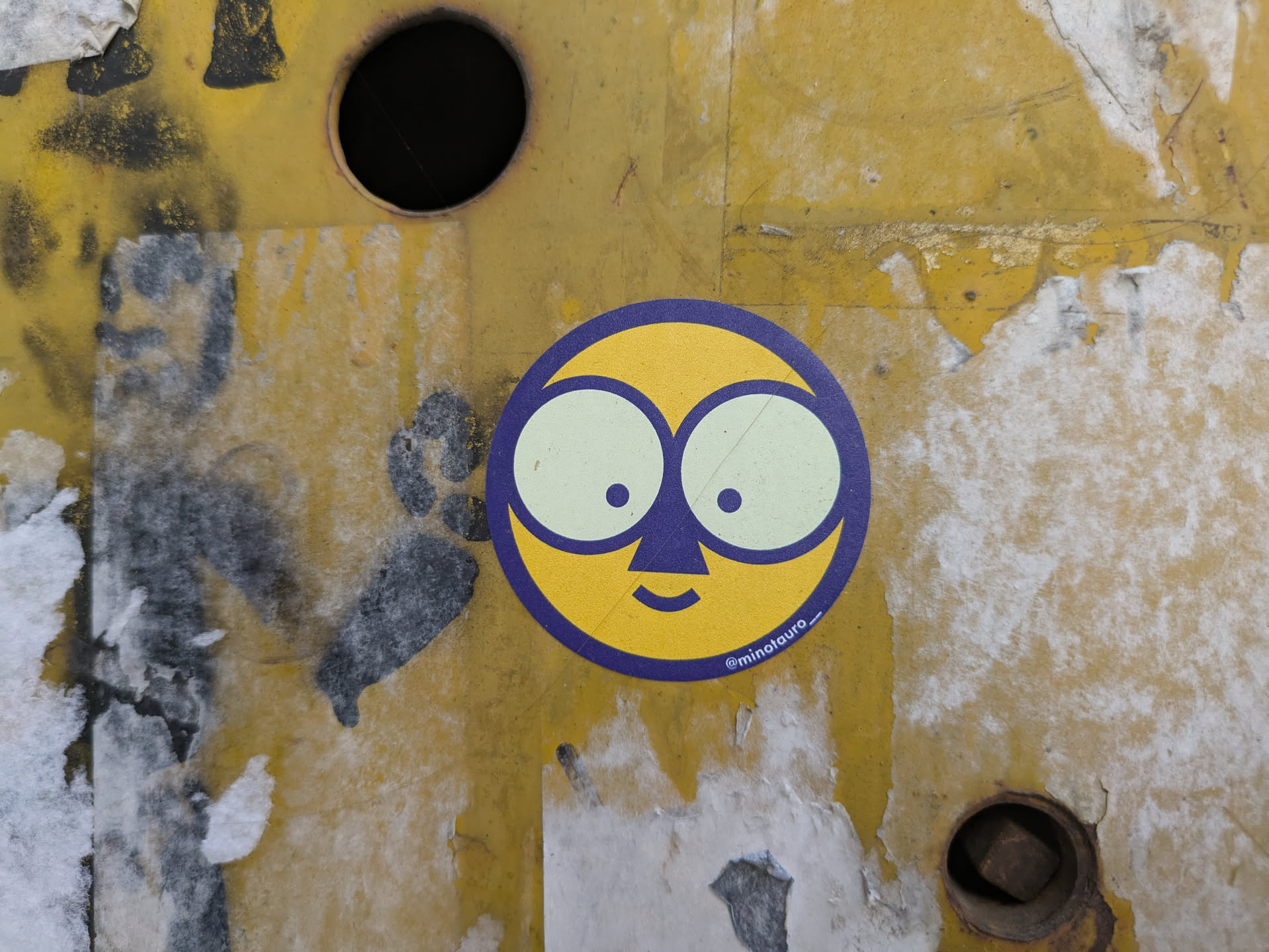 A round sticker features a yellow face with large, round, light green eyes and small black pupils. A dark blue nose and mouth complete the simple features. The face is outlined in dark blue. The sticker appears on a yellow, textured surface with some peeling paint.