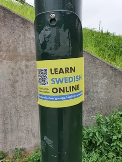 A yellow sticker on a green pole with a QR code and a Facebook group link to learn Swedish online.  The sticker encourages users to scan the QR code and join the online Swedish group.