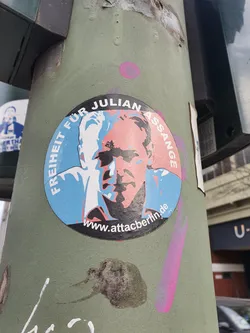 A sticker featuring a stylized portrait of Julian Assange with the text "FREIHEIT FÜR JULIAN ASSANGE" (Freedom for Julian Assange) and a website address. The sticker is affixed to a pole.