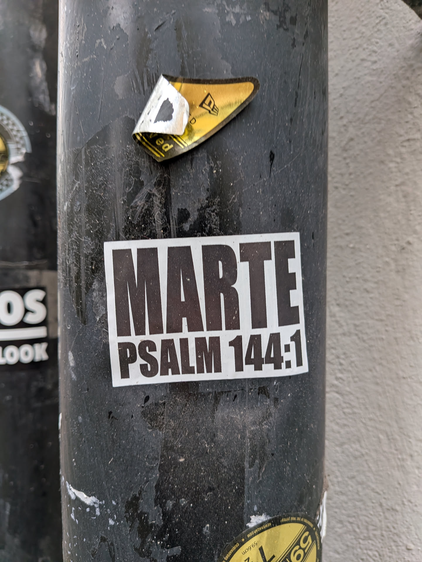 A rectangular sticker with a white background and dark, bold, sans-serif text. The top line prominently displays 'MARTE' in large uppercase letters, while the bottom line reads 'PSALM 144:1' in slightly smaller uppercase letters. The sticker is affixed to a dark, textured pole, showing some minor wear and tear.