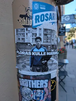 Street sticker A rectangular photographic sticker centered on a post. It portrays the character Saïd from the classic French film 'La Haine' standing with eyes closed in an urban housing block courtyard. While the background and jeans are in black and white, his zip-up track jacket has a pop of blue. Running horizontally across the center of his chest is a black banner containing bold white text that reads 'FLORAS KULLE MASSIVE'.