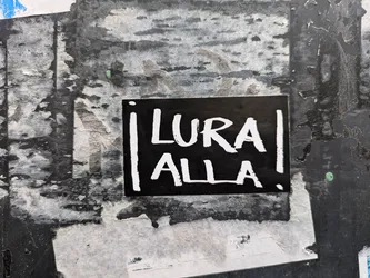 Black and white sticker with the text "Lura Alla" written in a stylized font. The sticker is attached to a textured surface with other layers of stickers and posters underneath.