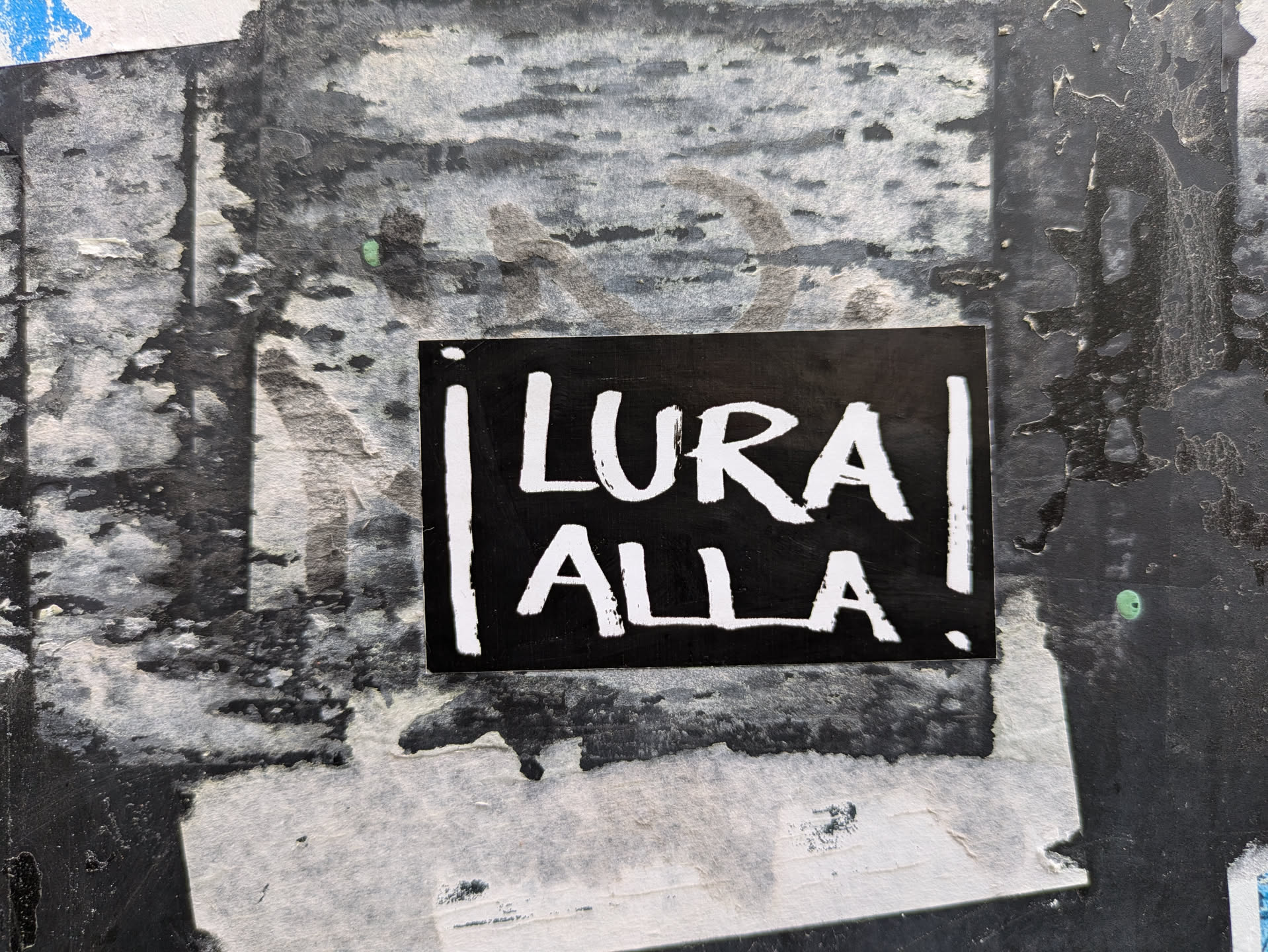 Black and white sticker with the text "Lura Alla" written in a stylized font. The sticker is attached to a textured surface with other layers of stickers and posters underneath.