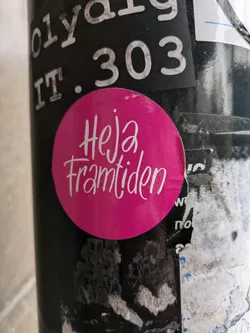 A pink round sticker with the text "Hej framtiden" written in a stylized white script.  The sticker is adhered to a surface with other stickers and markings.