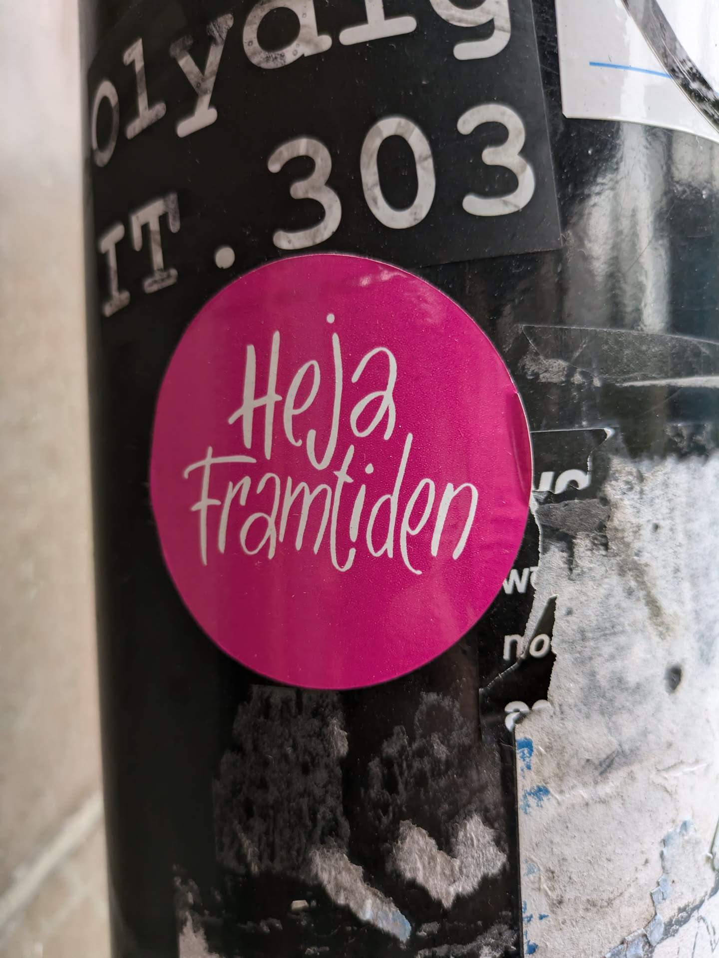 A pink round sticker with the text "Hej framtiden" written in a stylized white script.  The sticker is adhered to a surface with other stickers and markings.