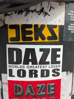 The image captures a close-up of multiple overlapping stickers adhered to a rough, dark surface, possibly a utility pole or wall. The central focus is a large white rectangular sticker with prominent black text. The top line reads 'DAZE', followed by smaller text 'WORLDS GREATEST LOVER', and then 'LORDS' in large, impactful letters. Directly above this white sticker is a smaller, bright yellow sticker with thick black letters spelling 'JEKS'. Below the main white sticker, a red sticker is partially visible, featuring a chain-link fence pattern and the word 'DAZE' in outlined black letters. The overall appearance suggests a collection of street art or urban tagging.
