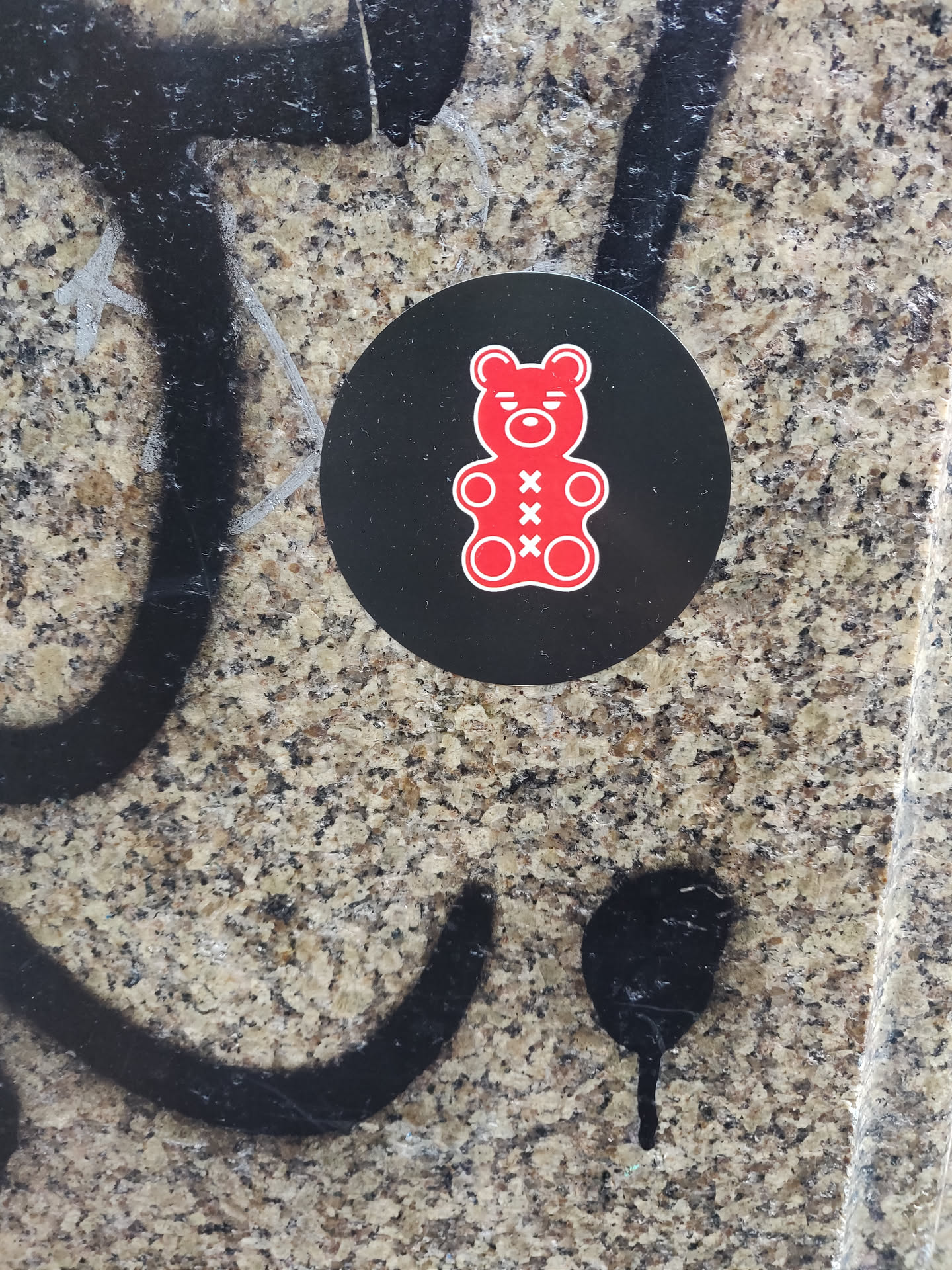A round black sticker with a red gummy bear in the center. The gummy bear has three 'x's on its belly. The sticker is placed on a granite surface with some graffiti around it.
