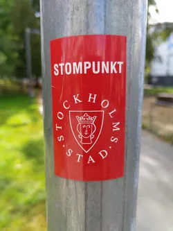 A red sticker on a metal pole with the text "STOMPUNKT" at the top and the Stockholm city logo below. The logo is a shield with a crowned female face inside.
