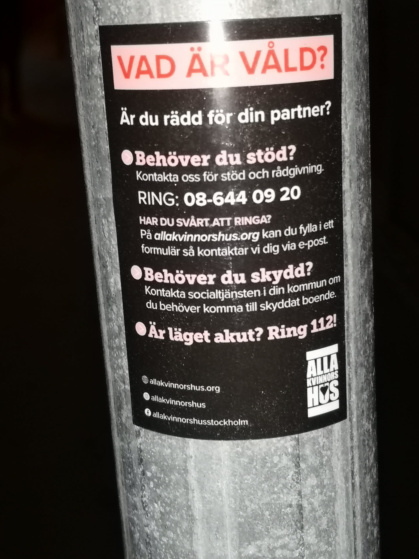 A rectangular, vertically oriented sticker with a black background, adhered to a metal pole. At the top, a light pink/white banner features the question 'VAD ÄR VÅLD?' (WHAT IS VIOLENCE?) in bold red text. Below this, the sticker presents information and questions in white text, such as 'Är du rädd för din partner?' (Are you afraid of your partner?), 'Behöver du stöd?' (Do you need support?), and 'Behöver du skydd?' (Do you need protection?). It provides a phone number '08-644 09 20', a website 'allakvinnorshus.org', and an emergency number 'Ring 112!'. The bottom left corner lists social media handles and a website, indicated by small icons (globe, Instagram, Facebook). In the bottom right, there is a white logo for 'ALLA KVINNORS HUS' (All Women's House) with stylized text.